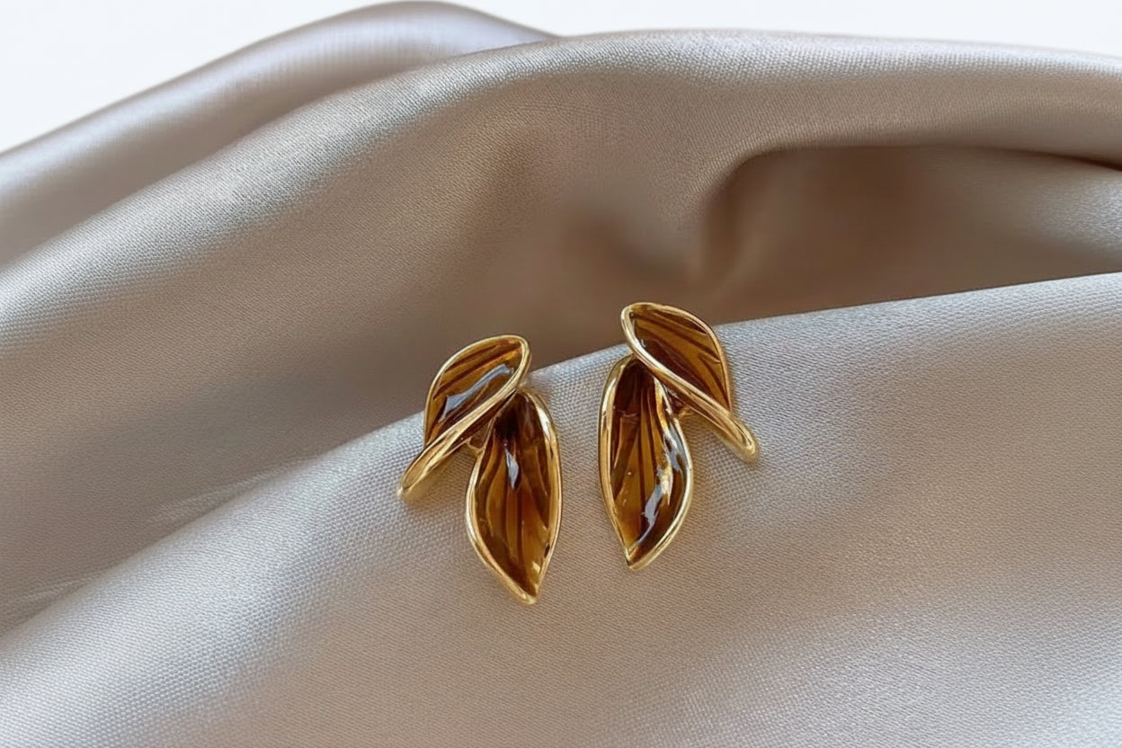 Gold leaf-shaped earrings with a brown painted gloss finish on a beige fabric background.