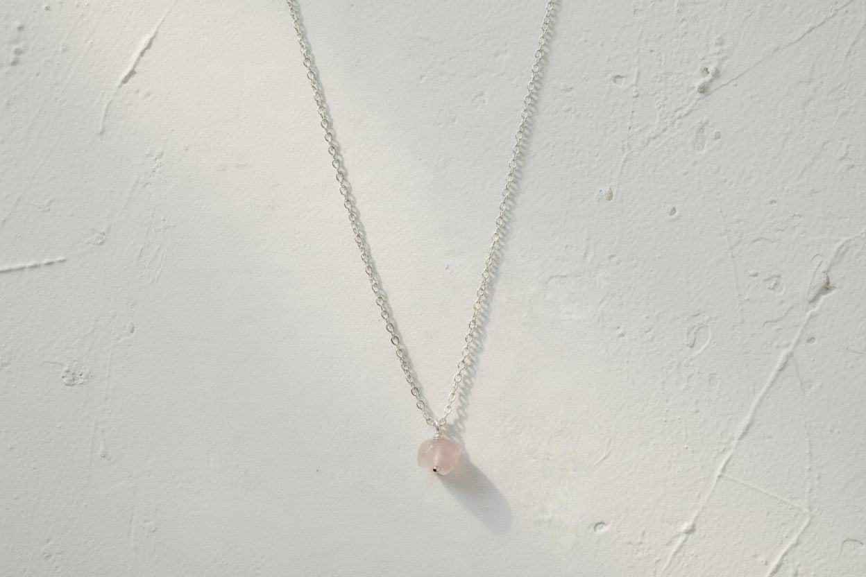 A silver necklace with a single rose quartz raw crystal pendant on a linen material.