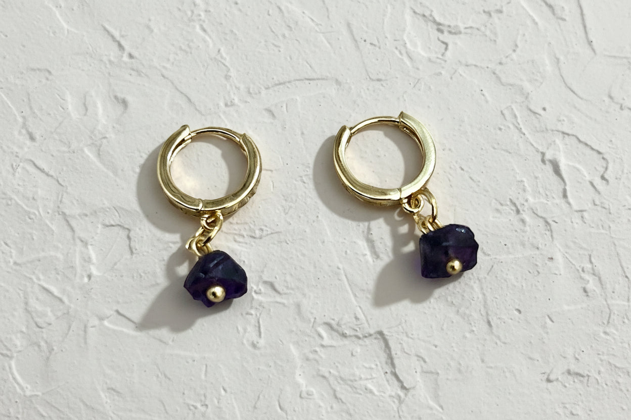 Gold hoop earrings with purple gemstones on a beige background.