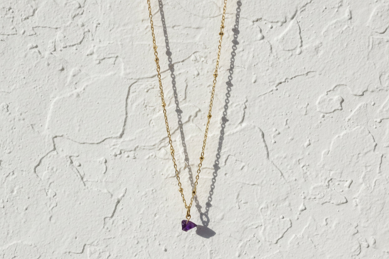Gold necklace with a purple raw amethyst crystal pendant on a white background.