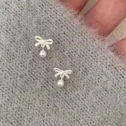 Pair of white bow earrings with pearls on a textured grey sleeve.