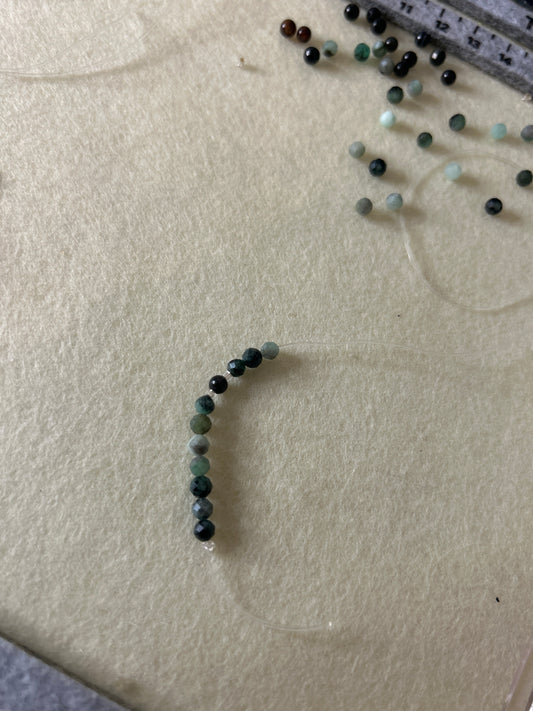 A few beads strung together to depict the start of a beaded necklace on a light fabric surface with a ruler for scale.