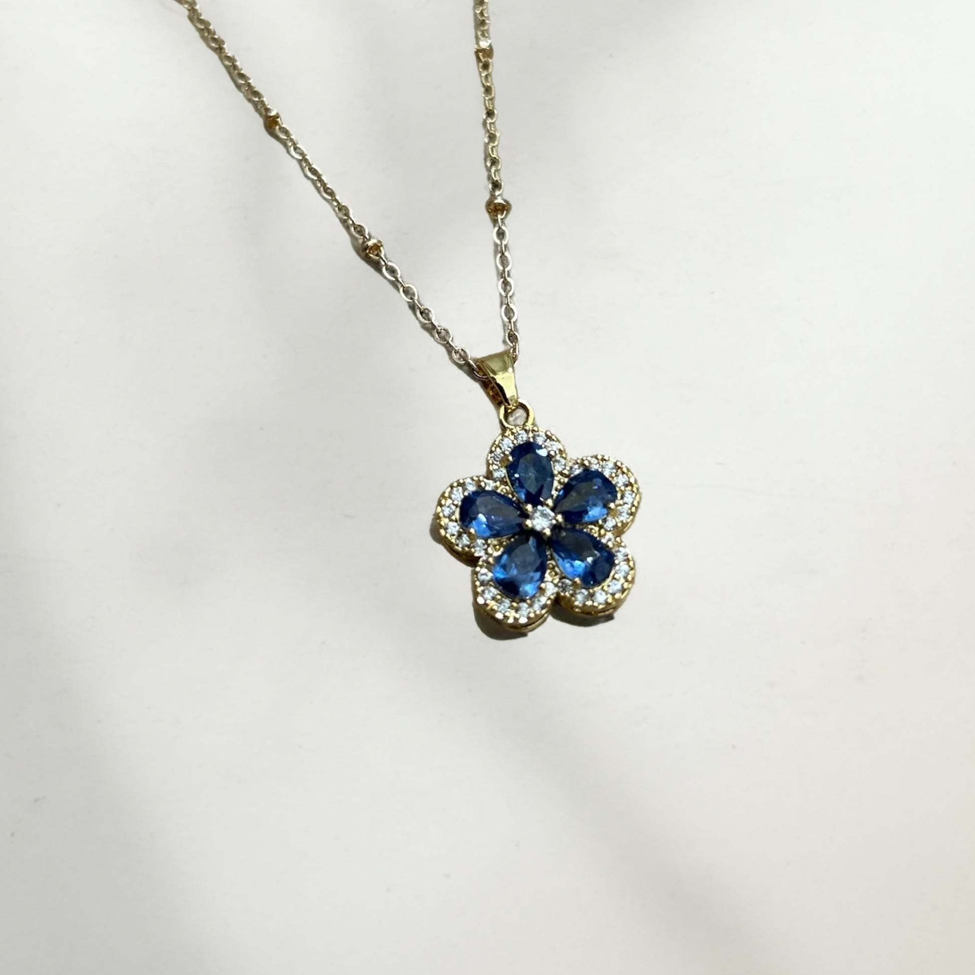 Flower-shaped pendant with blue crystals on a gold chain against a white background