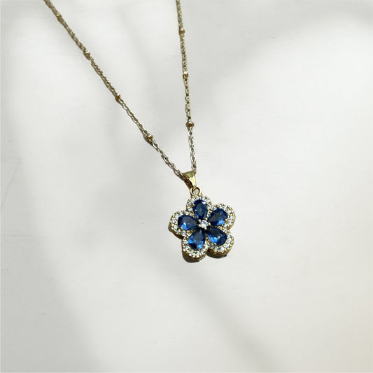Flower-shaped pendant with blue stones on a gold chain against a white background
