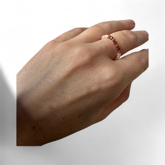 Hand wearing a bead ring with pearls, red and orange gemstones on a light grey background.